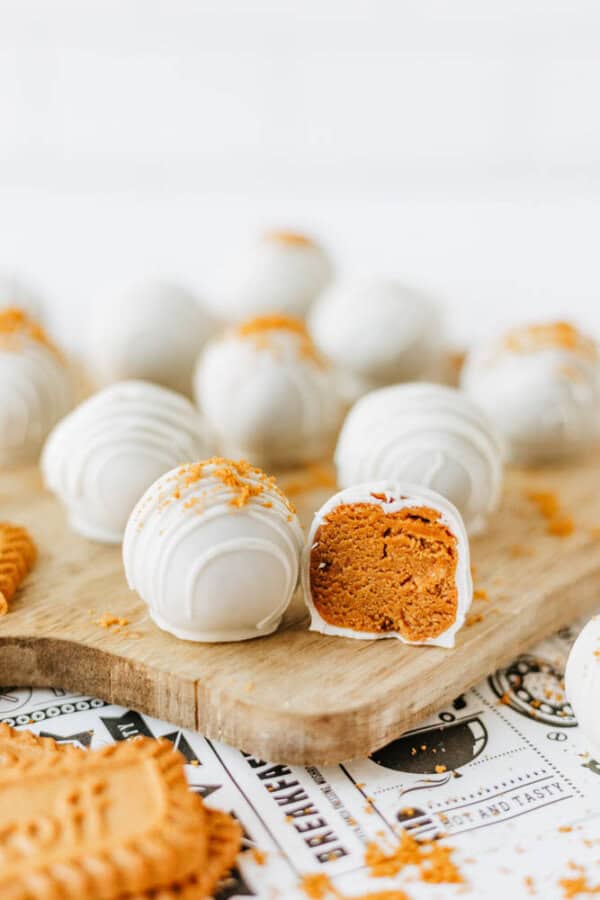 White chocolate-coated cookie truffles are arranged on a wooden board. One truffle is cut open, revealing a creamy orange-brown biscoff cookie butter delight filling. Crumbled cookie pieces are sprinkled on top of the truffles and board.
