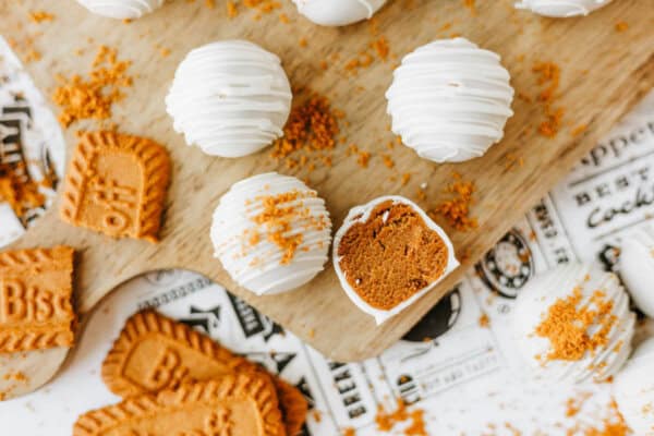 White chocolate-coated truffles, some whole and one cut in half to reveal a brown spiced filling, are arranged on a wooden board with crumbled Biscoff cookies—truly a biscoff cookie butter delight—plus whole cookies scattered around.