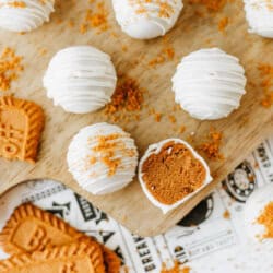 Round white chocolate truffles topped with crumbled cookies are arranged on a wooden board. One biscoff cookie butter delight truffle is cut open, revealing a rich brown filling and surrounded by Biscoff cookies and crumbs.