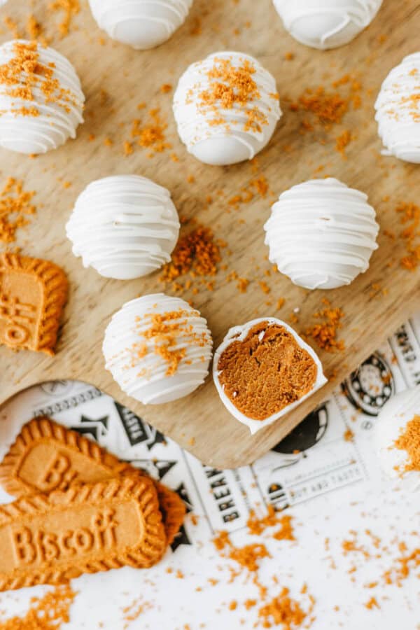 Round white chocolate truffles topped with crumbled cookies are arranged on a wooden board. One biscoff cookie butter delight truffle is cut open, revealing a rich brown filling and surrounded by Biscoff cookies and crumbs.