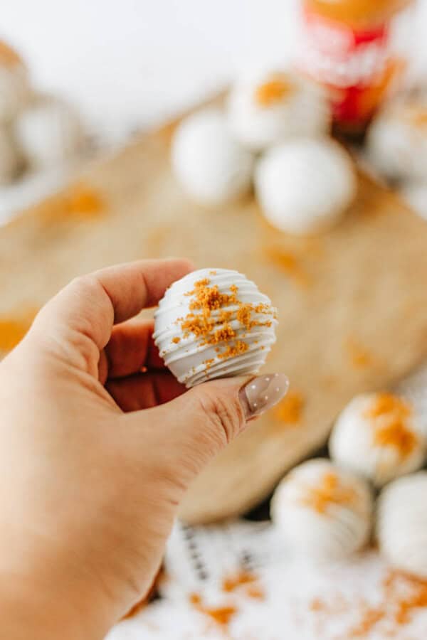 A hand holding a white chocolate-coated biscoff cookie butter delight topped with crumbled cookie pieces, with more similar treats and a wooden board in the blurred background.