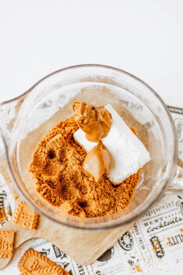 A glass mixing bowl containing crushed cookies, a block of cream cheese, and two spoonfuls of peanut butter creates a biscoff cookie butter delight, all placed on decorative paper with cookie pieces scattered around.