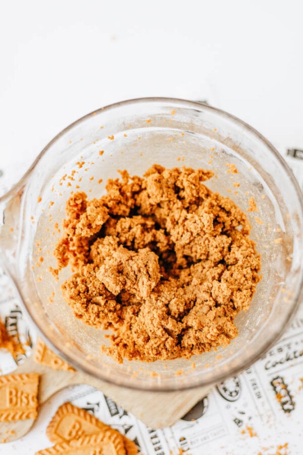 A glass mixing bowl containing a crumbly, brown biscoff cookie butter delight dough mixture sits on a patterned surface with cookie pieces nearby.