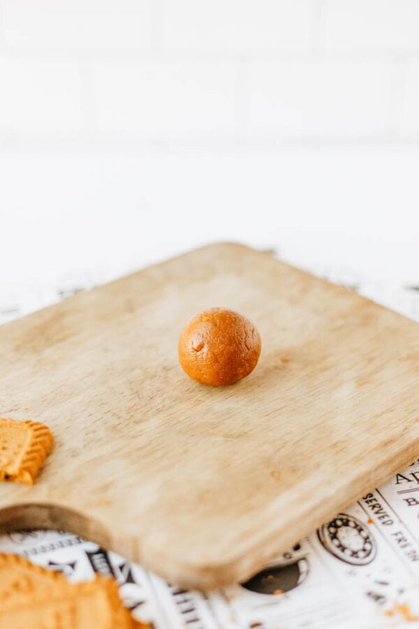 A single brown biscoff cookie butter delight dough ball sits on a wooden cutting board, with a partial biscuit visible in the bottom left corner. The background is white with scattered paper beneath the board.