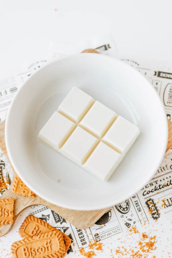 A white ceramic bowl contains six squares of white chocolate. Surrounding the bowl are Biscoff cookies and printed paper with decorative text, creating a biscoff cookie butter delight in a bright and clean setting.