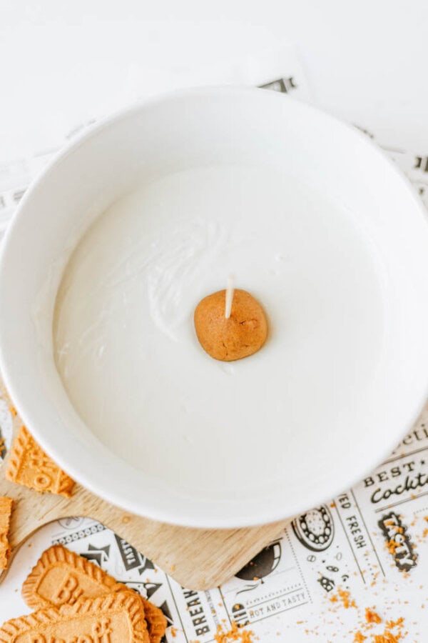 A round cookie on a stick is partially dipped in white melted chocolate, surrounded by biscoff cookie butter delight crumbles and paper on a light surface.