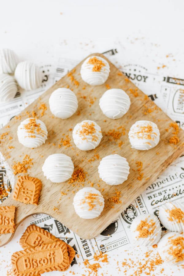 Nine white, round, textured cake balls are arranged on a wooden board, some topped with crumbled Biscoff cookies for a true biscoff cookie butter delight. Extra crumbled and whole cookies are scattered on the white surface.