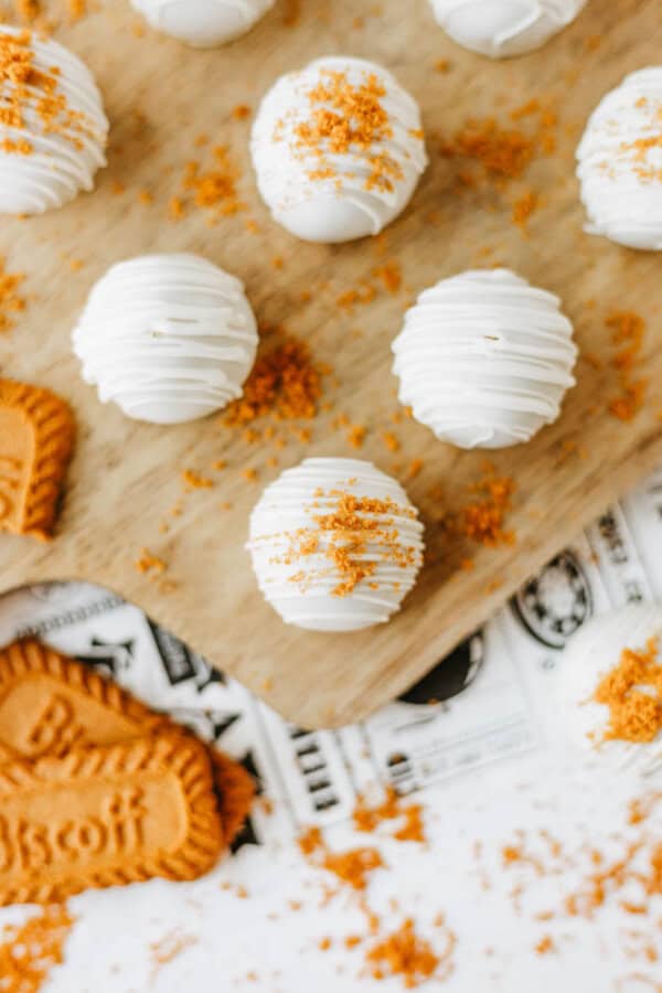 A wooden board with round white chocolate truffles, some topped with crumbled caramel cookies for a biscoff cookie butter delight. Cookie crumbs and whole caramel cookies are scattered on the board and printed paper below.