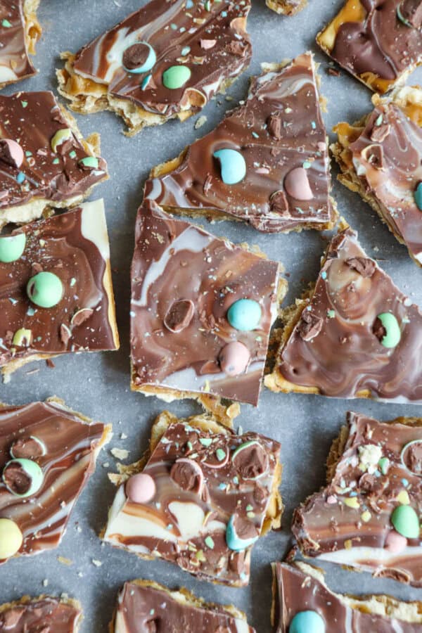 Pieces of swirled milk and white chocolate bark topped with pastel-colored candy-coated chocolate eggs, scattered on a sheet of parchment paper.