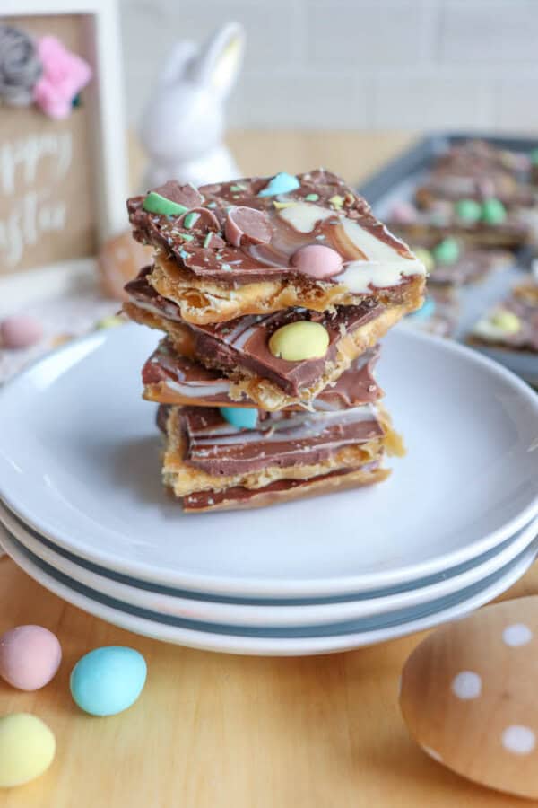 A stack of chocolate bark pieces topped with pastel candy eggs sits on a stack of white plates. More bark and candies are in the background, along with an Easter bunny figurine and festive decor.