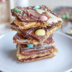 A stack of chocolate toffee bars topped with colorful candy-coated chocolates and white chocolate swirls sits on a white plate, with a blurred background.