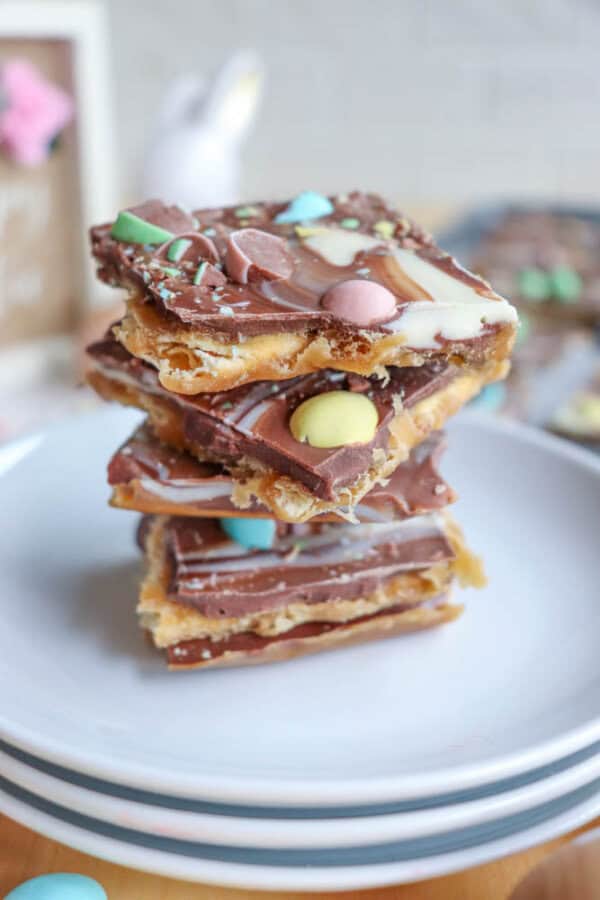 A stack of chocolate toffee bars topped with colorful candy-coated chocolates and white chocolate swirls sits on a white plate, with a blurred background.