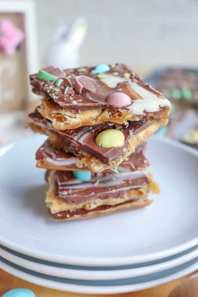 A stack of chocolate toffee bars topped with colorful candy-coated chocolates and white chocolate swirls sits on a white plate, with a blurred background.