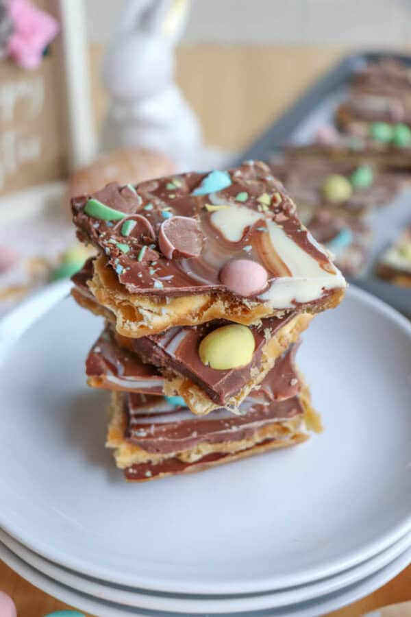 A stack of chocolate bark pieces topped with colorful candy eggs and drizzled with white and milk chocolate, placed on a white plate. Easter-themed decorations are blurred in the background.