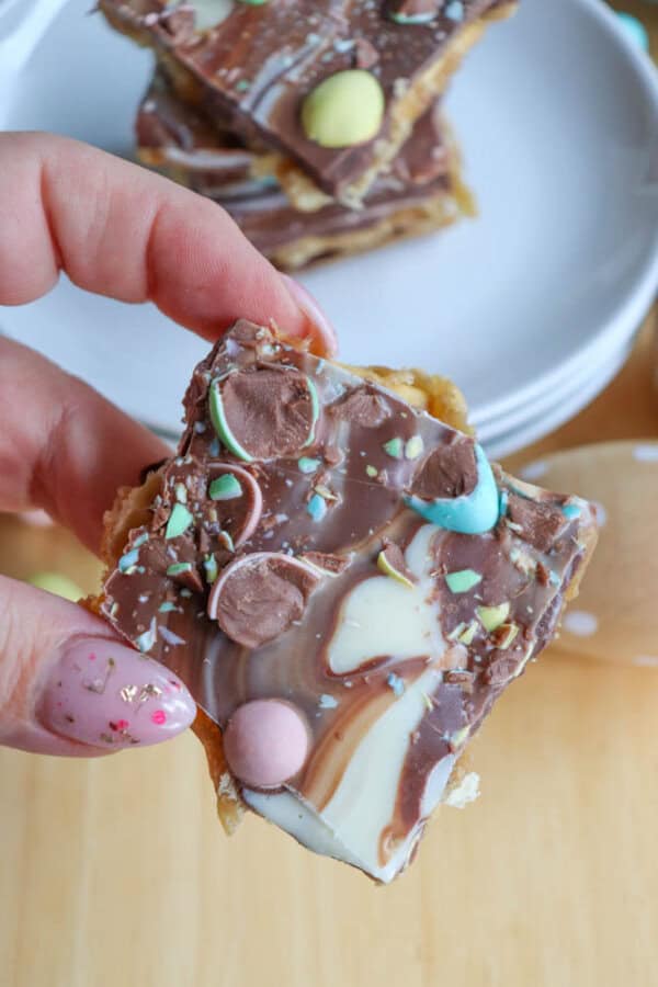 A hand holding a square piece of chocolate bark topped with colorful candy pieces and swirls of white and milk chocolate, with a stack of plates in the background.