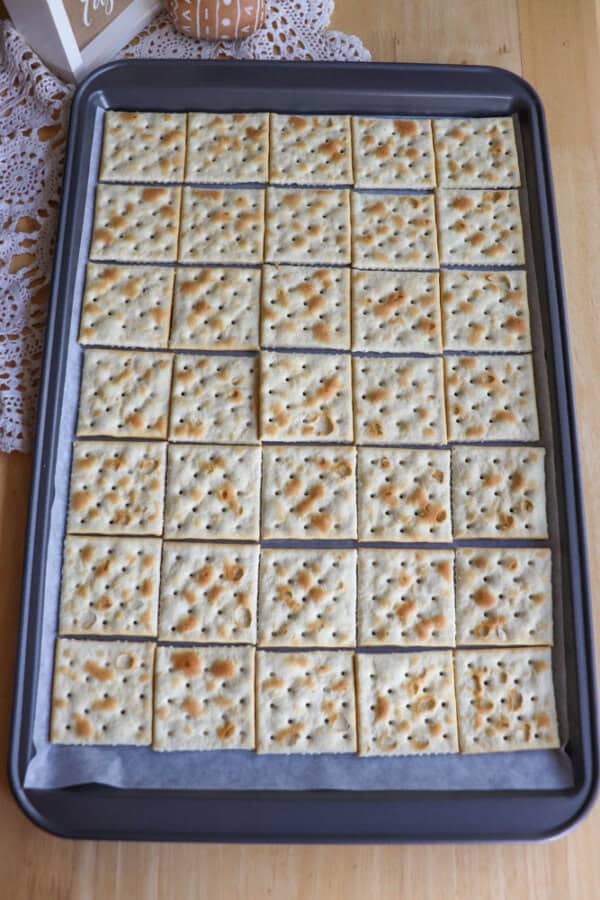 A baking sheet lined with parchment paper holds 40 evenly arranged saltine crackers in a 5 by 8 grid on a wooden table.