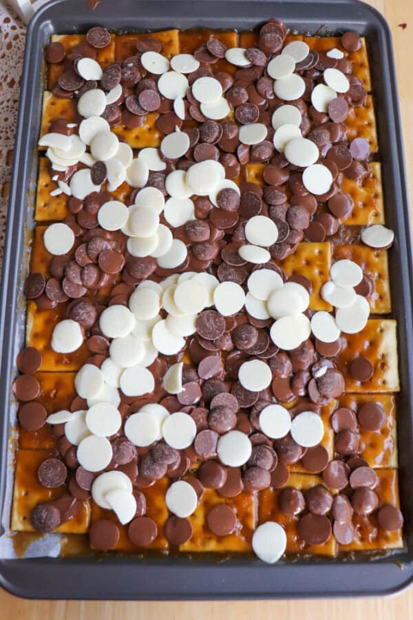 A baking sheet filled with saltine crackers topped with caramel sauce and a mix of milk and white chocolate melting wafers, ready to be baked.