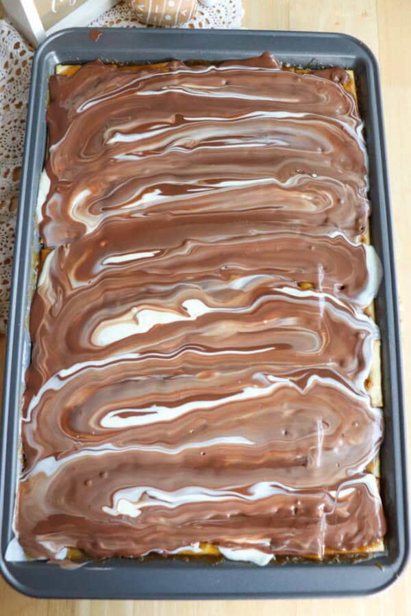 A rectangular baking pan filled with dessert bars topped with swirled layers of milk chocolate and white cream, creating a marbled pattern on the surface. The pan is resting on a wooden surface.