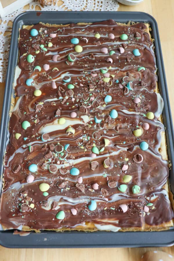 A sheet pan covered with a layer of swirled chocolate and white chocolate, topped with colorful candy-coated eggs, chocolate pieces, and sprinkles, resembling a festive dessert bark.