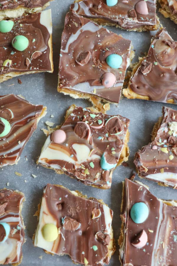 Squares of marbled milk and white chocolate bark with pastel candy-coated chocolates and crushed candy pieces scattered on top, arranged on parchment paper.