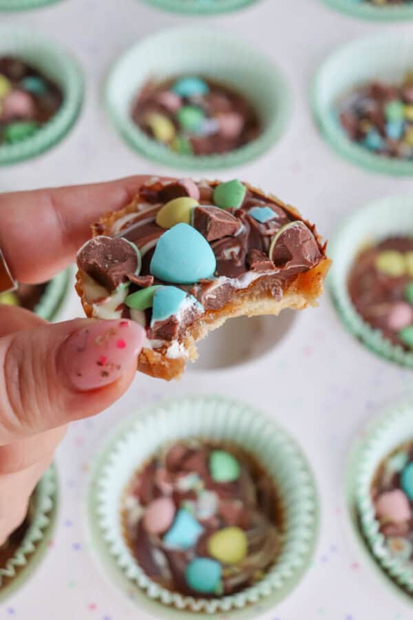 A hand with painted nails holds a partially eaten chocolate treat topped with colorful candy pieces, with more similar treats in green cupcake liners visible in the background.