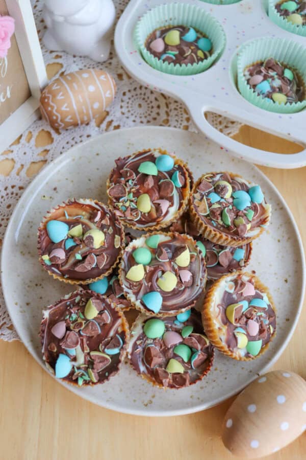 A plate of cupcakes topped with chocolate and colorful candy eggs sits on a wooden table, surrounded by decorated Easter eggs and a muffin tray with more cupcakes in pastel liners.