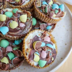 A plate of cupcake-sized chocolate treats topped with swirled white and milk chocolate and colorful candy-coated chocolate eggs, arranged on a light-colored speckled plate.
