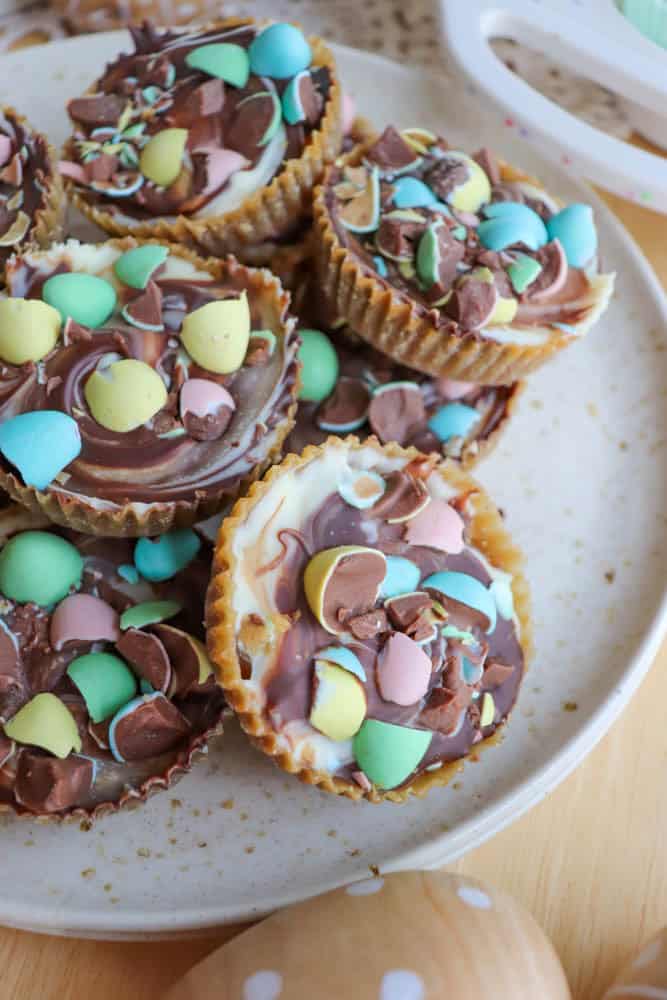 A plate of cupcake-sized chocolate treats topped with swirled white and milk chocolate and colorful candy-coated chocolate eggs, arranged on a light-colored speckled plate.