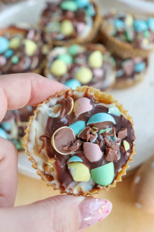 A hand holds a chocolate cup topped with crushed pastel-colored candy eggs. More similar chocolate treats are on a tray in the background. The person has a pink and white patterned manicure.