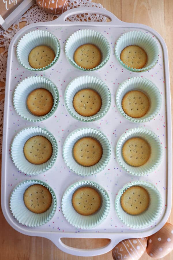 A white muffin tray with twelve slots holds paper liners, each containing a single round vanilla wafer cookie. The tray is on a wooden surface with a lace doily and decorative eggs nearby.