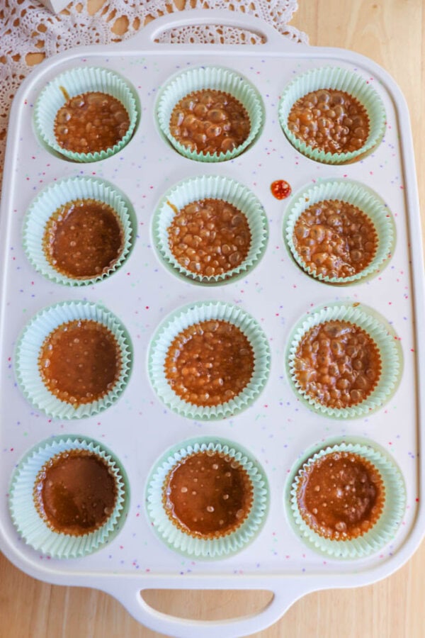 A muffin tin lined with light green paper cups, each filled with a bubbly caramel-like mixture, sits on a wooden surface near a lace doily.
