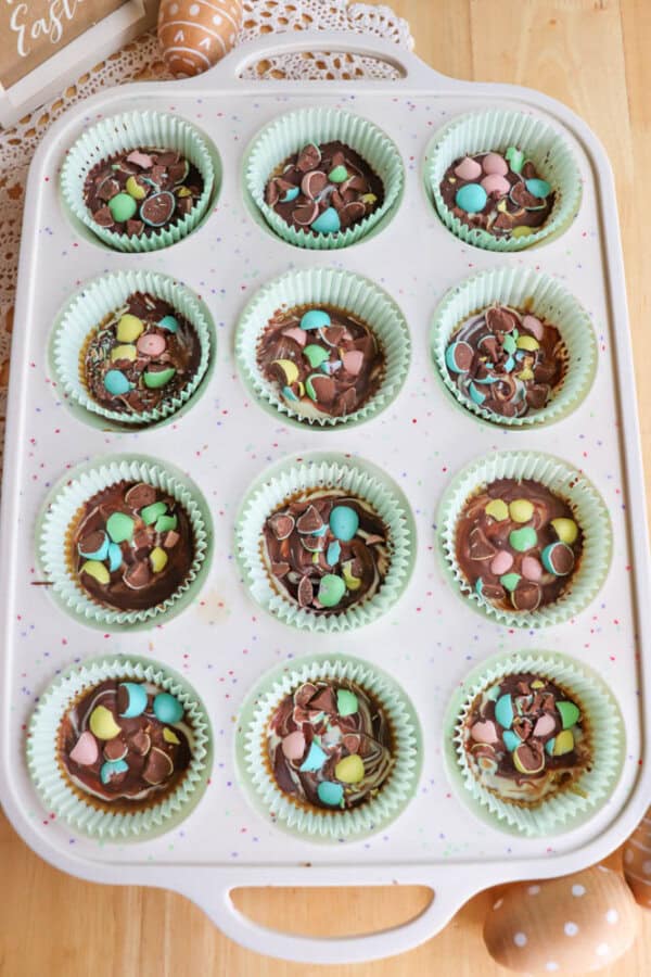 A white muffin tray holds 12 cupcakes in pastel green liners, each topped with chocolate and colorful candy eggs. The tray rests on a wooden table decorated with Easter-themed items.