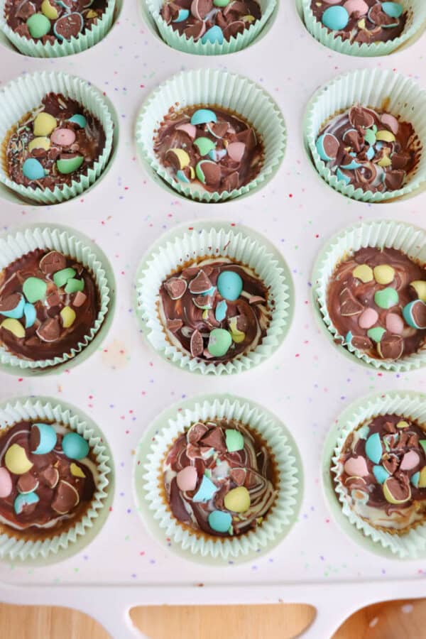 A muffin tin filled with cupcakes in pale green paper liners, topped with chocolate frosting and colorful pastel candy pieces, viewed from above.