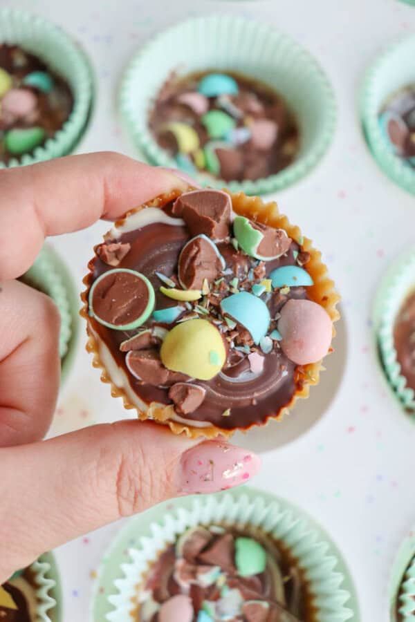 A hand holding a chocolate cup topped with crushed pastel candy eggs, surrounded by more chocolate cups in green paper liners.