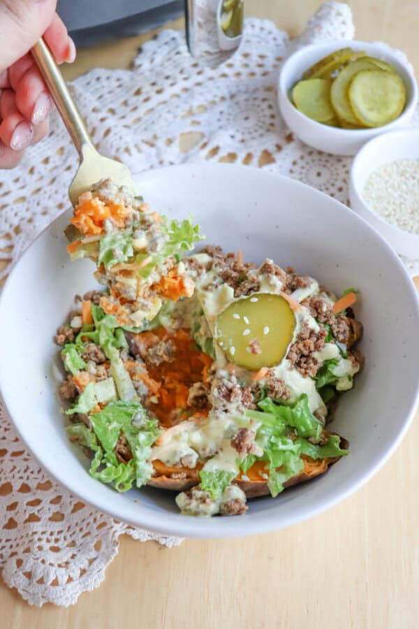 A white bowl filled with a salad of ground meat, lettuce, shredded carrots, pickles, and creamy dressing. A fork is lifting a bite from the bowl. Two small dishes of pickles and sesame seeds are in the background.