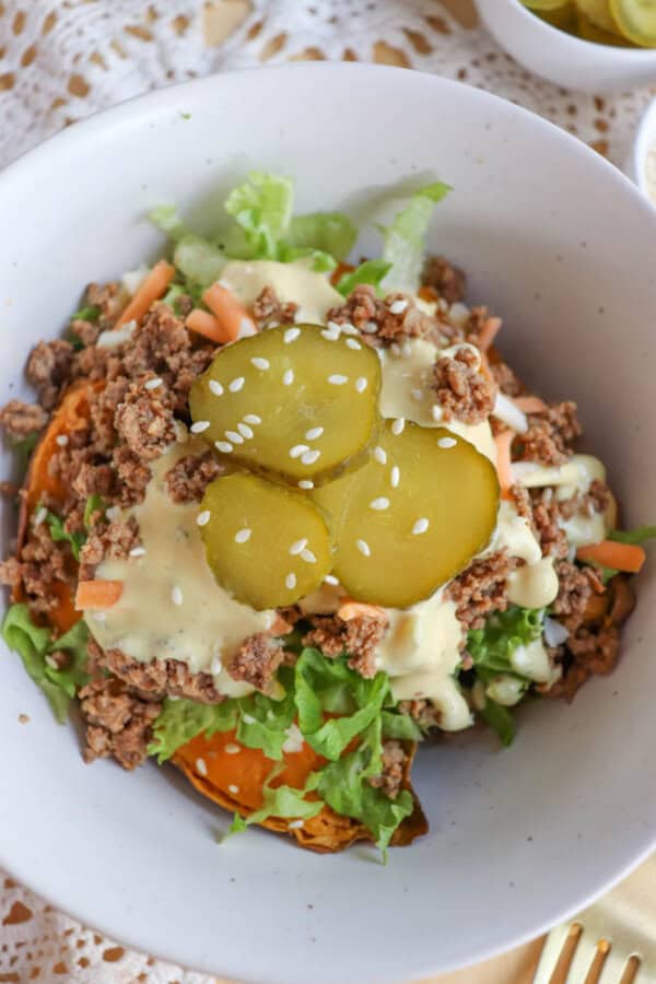 A bowl of salad topped with ground beef, shredded lettuce, sliced pickles, shredded carrots, creamy dressing, and a sprinkle of sesame seeds. The salad is served in a white bowl on a lace-patterned tablecloth.