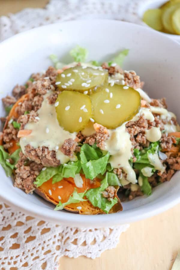 A bowl filled with ground beef, lettuce, sweet potato slices, sliced pickles, creamy dressing, and sprinkled with sesame seeds, set on a lace tablecloth.