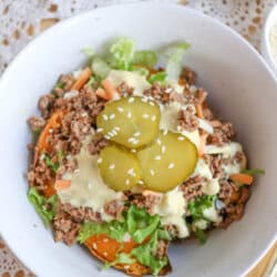 A white bowl filled with ground beef, shredded lettuce, carrots, sauce, and topped with sesame seeds and sliced pickles. A fork and a side dish of pickles are placed nearby on a lace tablecloth.