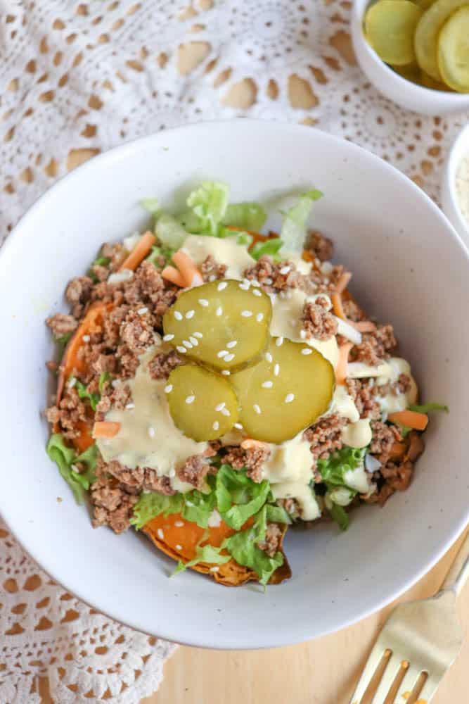 A white bowl filled with ground beef, shredded lettuce, carrots, sauce, and topped with sesame seeds and sliced pickles. A fork and a side dish of pickles are placed nearby on a lace tablecloth.