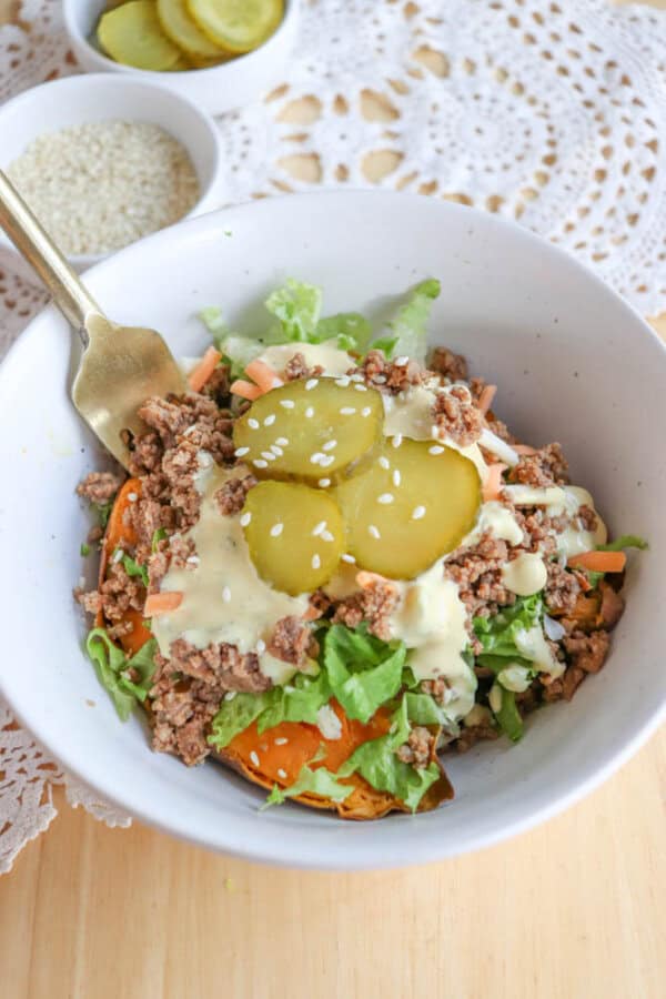 A white bowl filled with ground beef, shredded lettuce, sliced pickles, shredded carrots, and a creamy dressing, topped with sesame seeds. A fork rests inside the bowl; pickles and sesame seeds are in small bowls nearby.