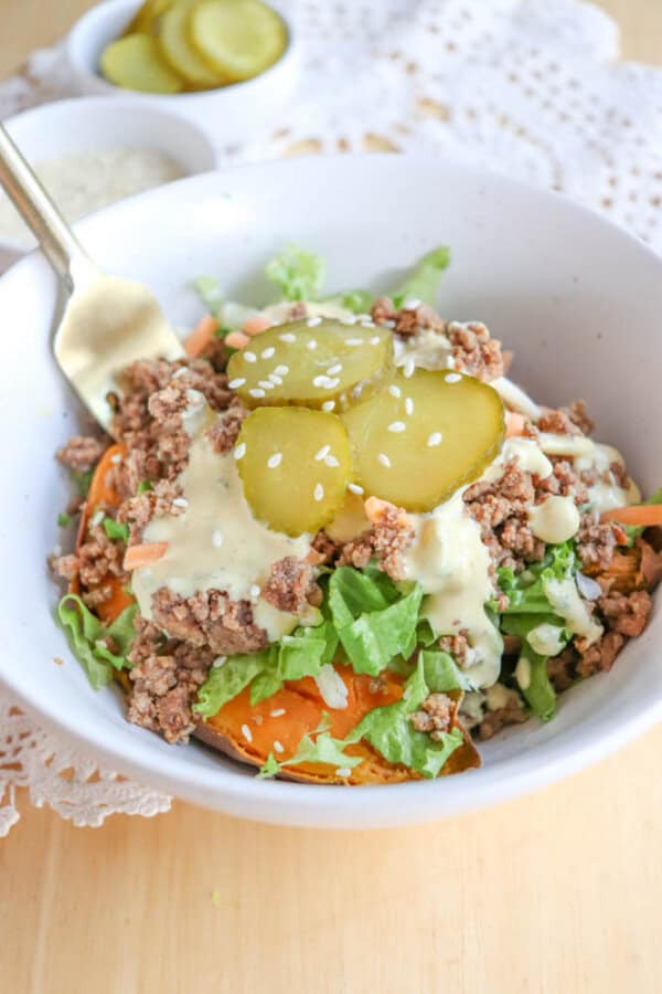 A bowl filled with ground meat, shredded lettuce, sliced pickles, and creamy dressing, topped with sesame seeds, sits on a lacy cloth with a fork. A small dish of pickles is in the background.