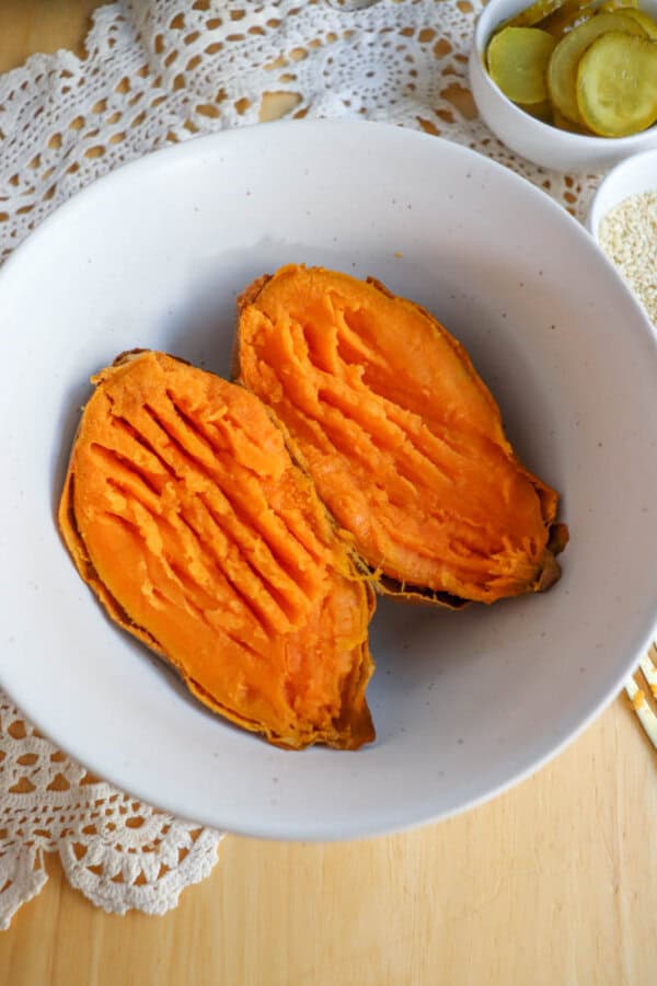 Two halves of a baked sweet potato with ridges on the flesh sit in a white bowl, on a wooden table with a lace doily. In the background, a small bowl of sliced pickles is partially visible.