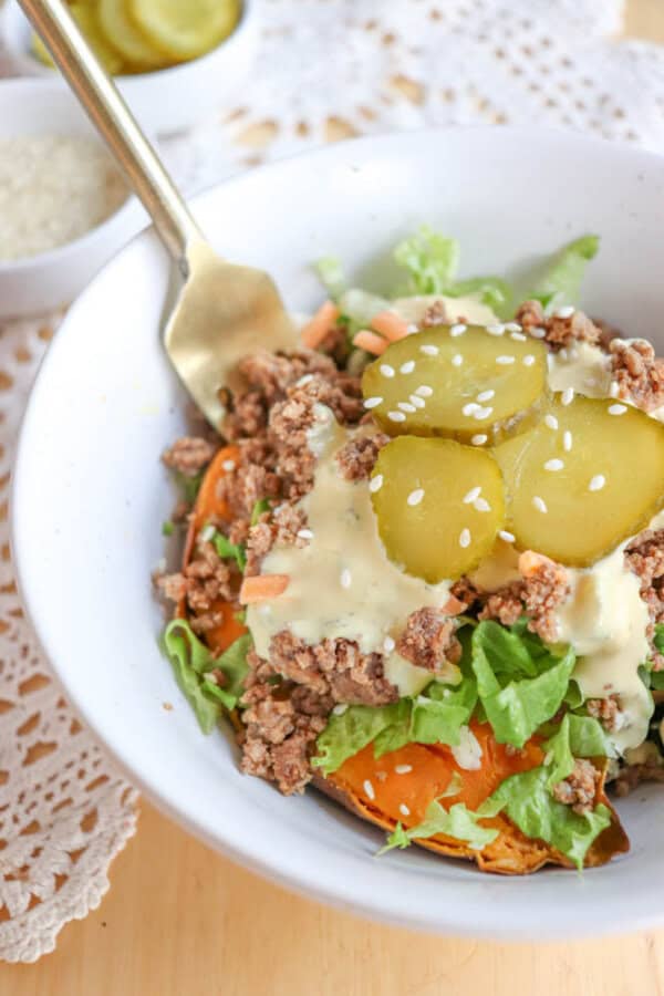 A white bowl filled with lettuce, ground meat, sliced pickles, shredded carrots, sesame seeds, and a creamy dressing, with a gold fork resting on the side. A doily and small bowls are in the background.