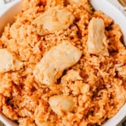 A bowl of orange-colored chicken and rice is shown close up, with chunks of chicken mixed into the rice. A fork and spoon are placed beside the bowl. Text above reads Sweet & Savory One Pot Chicken.