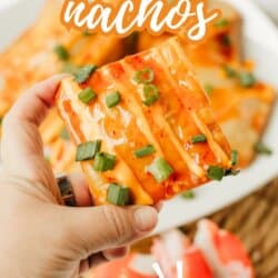 A hand holds a crispy crab rangoon nacho topped with orange sauce and chopped green onions. More nachos and pieces of imitation crab are seen on a plate in the background. Text reads: “Crab Rangoon Nachos, so good!”.