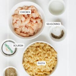 Overhead view of labeled ingredients in bowls: diced chicken, rotini pasta, Boursin cheese, grated cheese, seasoning, salt, pepper, and a small bowl of liquid, all arranged on a white surface.