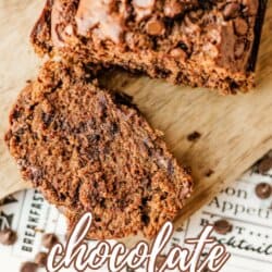 A loaf of chocolate zucchini bread on a wooden cutting board with one slice cut off. Text on the image reads “chocolate zucchini bread.” Chocolate chips are sprinkled on top and around the bread.