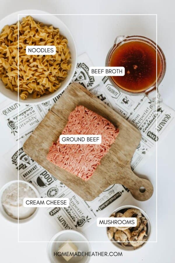 Flat lay of ingredients for a recipe: uncooked egg noodles, a glass of beef broth, raw ground beef on a wooden board, a container of cream cheese, and a bowl of mushrooms, all labeled.
