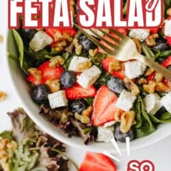 A colorful strawberry feta salad with spinach, blueberries, walnuts, and feta cheese cubes in a white bowl, topped with fresh strawberries. A fork rests on the salad. Text over the image reads: Strawberry Feta Salad, SO GOOD.