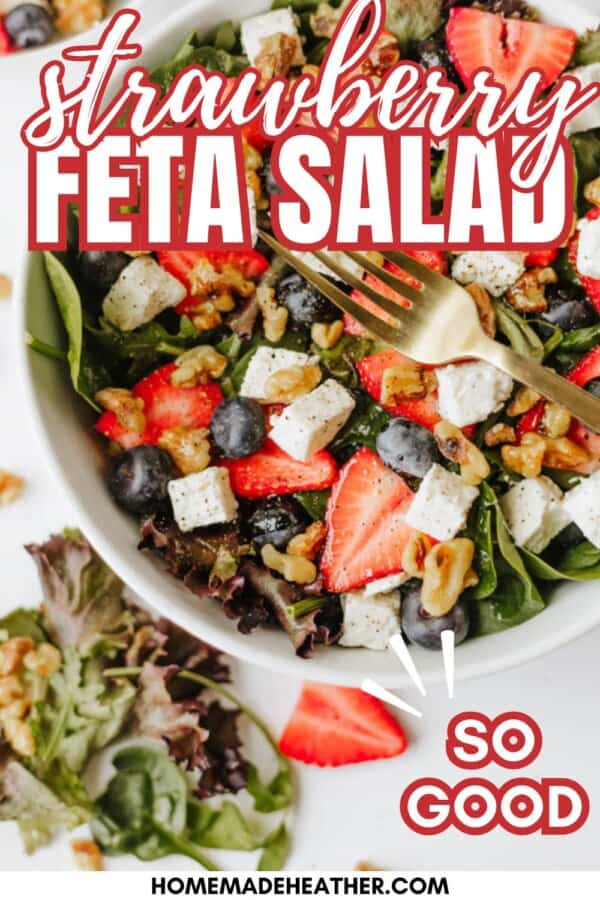 A colorful strawberry feta salad with spinach, blueberries, walnuts, and feta cheese cubes in a white bowl, topped with fresh strawberries. A fork rests on the salad. Text over the image reads: Strawberry Feta Salad, SO GOOD.